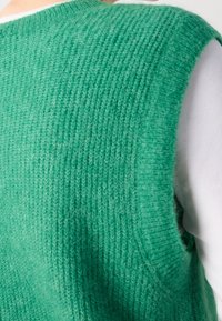 Veste sans manches verte en maille avec une finition texturée, dotée d'un col rond et d'une chemise lisse de couleur claire en dessous.