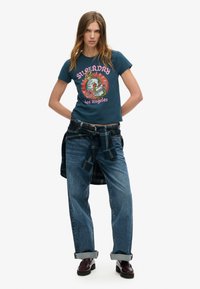 Blauwe katoenen t-shirt met een afbeelding van een slang en rozen, gecombineerd met losse spijkerbroek en een geruit overhemd om de taille geknoopt.