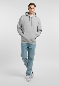 Felpa grigia chiara con cappuccio e tasca frontale, abbinata a jeans azzurri chiari e sneakers bianche. Tessuto casual e vestibilità comoda.