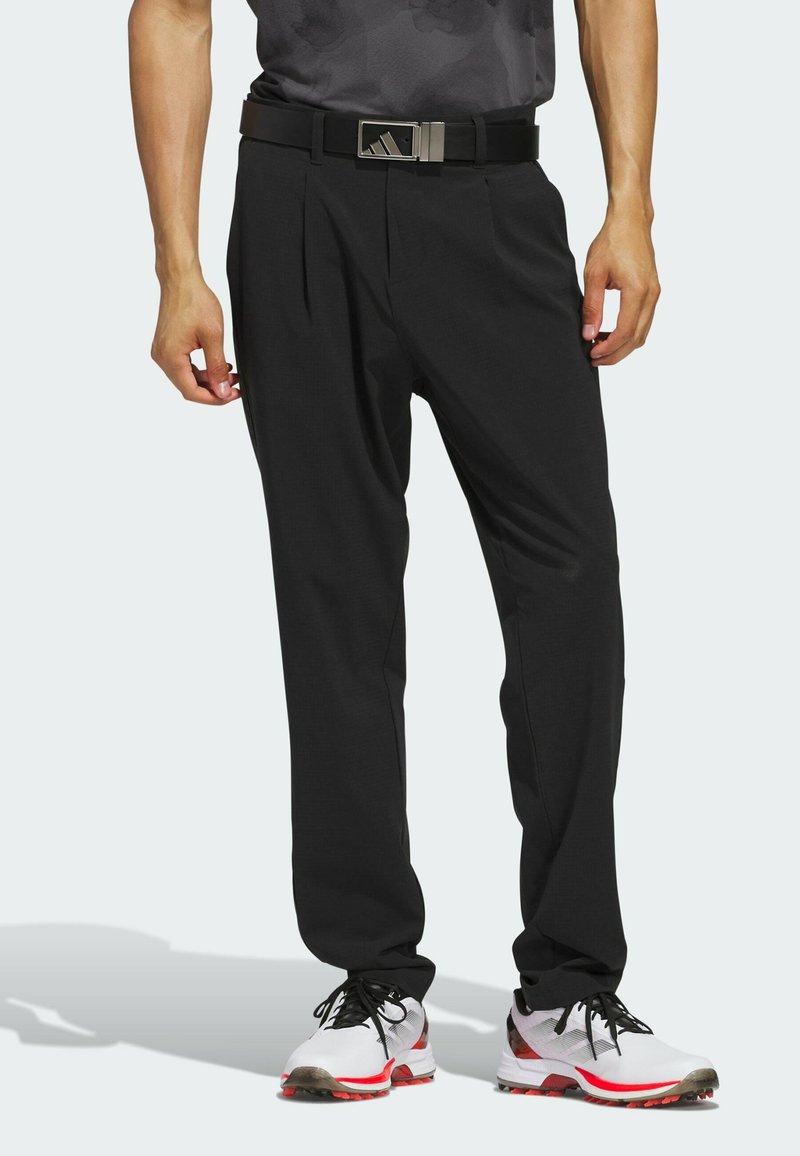 Pantalones deportivos negros con pliegues y corte recto. Llevados con un cinturón negro que cuenta con una hebilla metálica. Combinados con zapatos blancos y rojos.