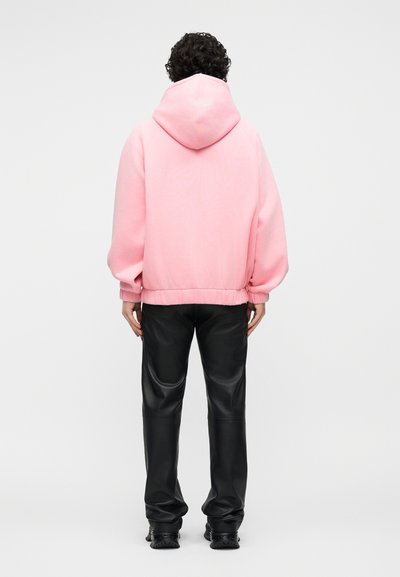 Personne aux cheveux bouclés portant un sweat à capuche rose ample avec la capuche relevée, un pantalon en cuir noir et des chaussures noires, debout de dos.