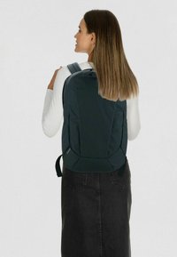 Dunkelgrüner Rucksack mit glatter, strukturierter Oberfläche, ausgestattet mit gepolsterten Trägern und einem Reißverschlussfach. Das Modell trägt ein weißes Oberteil und einen schwarzen Rock.