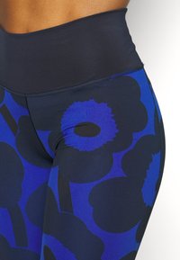Detailní záběr osoby s vysokými legínami, na kterých je výrazný tmavě modrý a černý květinový vzor na bílém pozadí.