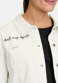 Veste en denim blanc avec un texte brodé "lovely sunny days" en noir, et trois cœurs dorés sur la poche poitrine. Boutons en métal argenté.