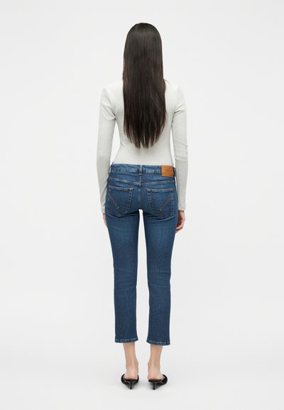 Femme aux longs cheveux foncés portant un haut gris clair à manches longues, un jean bleu court et des mules noires à talons, debout, de dos.