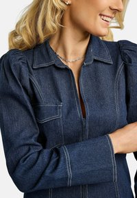 Traper košulja tamnoplave boje s izrađenim krojem, napuhanim rukavima i kontrastnim bijelim šavovima. Ima prednji džep i patentni zatvarač.