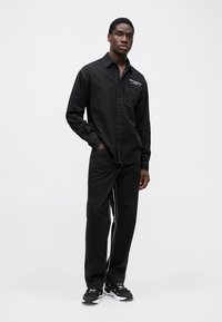 Camisa negra de botones con un bolsillo en el pecho, combinada con pantalones negros que tienen franjas laterales blancas. El modelo lleva zapatillas negras.