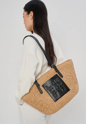 Vrouw met lang donker haar draagt een grote gevlochten lichtbruine handtas met een zwarte leren patch en bandjes over haar schouder.