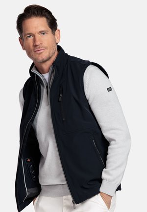 Man draagt een lichtgrijze trui onder een mouwloos zwart vest met ritssluiting, poseert met handen in de zakken van zijn witte broek tegen een witte achtergrond.