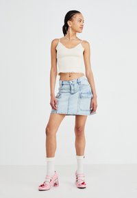Lichtgekleurde cropped tanktop met golvende randen, gecombineerd met een lichtgekleurde denim rok met grote zakken en roze blokhakken.