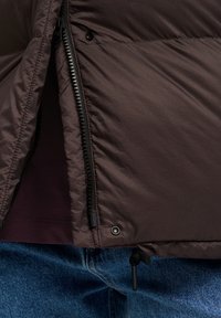 Veste isolée marron avec une coque texturée, dotée de fermetures éclair et de détails à pression, superposée sur une chemise violette, accompagnée de jeans bleus.