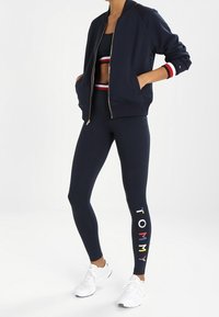 Námořnická bomber bunda na zip, přiléhavé námořnické legíny s vícebarevným nápisem „TOMMY“ po straně, doplněné bílými teniskami.