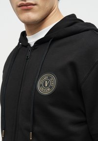 Άνδρας που φοράει μαύρο φούτερ με φερμουάρ, με κυκλικό χρυσό και μαύρο λογότυπο Versace Jeans Couture στο στήθος, πάνω από ένα λευκό μπλουζάκι με στρογγυλή λαιμόκοψη.