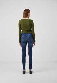 Oliven langermet topp kombinert med mørkeblå skinny-jeans med frynsete kant og baklommer, vist fra baksiden.