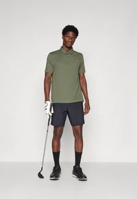 Σκούρα πράσινο πόλο μπλουζάκι με κοντά μανίκια, μαύρο σορτς, μαύρες κάλτσες και παπούτσια γκολφ. Κρατάει μπαστούνι γκολφ, φοράει λευκό γάντι γκολφ.