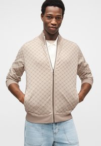 Veste bombardier beige clair avec un motif texturé en relief, dotée d'une fermeture éclair à l'avant, de poignets côtelés et de poches. Portée sur un polo blanc.