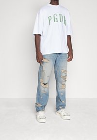 Vit kortärmad t-shirt med grön "PGDA" text; ljusblå slitna jeans med flera revor; vita sneakers. Enkelt och avslappnat utseende.