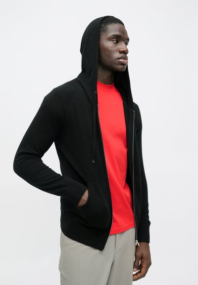 Homem a usar um hoodie preto com fecho e capuz puxado, camisa vermelha e calças cinza claro, em pé com uma mão no bolso contra um fundo branco.