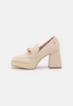 Mocasines de tacón alto en color beige con un acabado texturizado. Presentan un tacón ancho, una correa acolchada con detalles en tono dorado y una puntera redondeada.