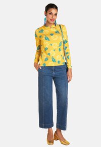 Gelbes Langarmshirt mit Blumenmuster, kombiniert mit weit geschnittenen Jeans. Akzentuiert mit goldenen High Heels und hängenden blauen Ohrringen.
