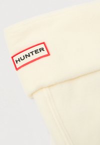 Hvit fleece-stoff med mansjett, med en rektangulær logo-patch som har "HUNTER" i svarte bokstaver med rød kant. Glatt tekstur.