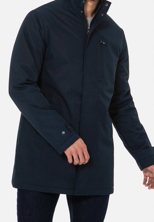 Homme portant un manteau d'hiver isolé bleu marine avec un col montant, une poche poitrine zippée et un revers à bouton-pression sur la manche gauche.