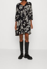 Robe noire à fleurs avec des manches longues et un décolleté en v, ornée de motifs floraux blancs. Associée à des bottes en cuir noires montantes.