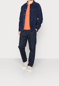 Giacca di velluto a coste blu navy sopra una maglietta arancione, abbinata a pantaloni scuri e sneakers beige chiaro, con chiusura a bottone e tasca laterale.