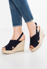 Mörkblå mocka kilklacksandaler med korsade remmar, öppen tå och vävd jute-baserad sula. Justeras med en spännrämsrem bak.