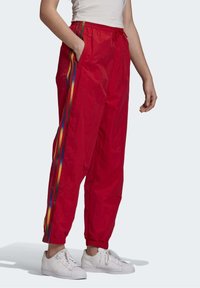 Červené sportovní kalhoty vyrobené z lehkého materiálu, s duhově pruhovanými bočními částmi, elastickými manžetami a páskem na stažení.