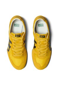 Žluté semišové tenisky s černými pruhy, šněrovací design, texturovaný svršek a logo na jazyku. Plátová gumová podrážka pro trakci.