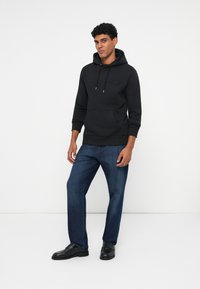Sudadera de algodón negra con bolsillo frontal y cordones ajustables, combinada con jeans de pierna recta azules y zapatos de cuero negros.