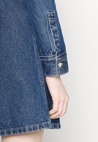 Sukienka dżinsowa w ciemnoniebieskim kolorze, z długimi rękawami zakończonymi mankietami na guziki oraz prostym krojem. Wyraźne boczne rozcięcie dla swobody ruchu.