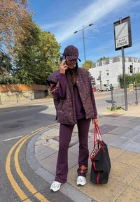 Fialová vetrová vesta so vzorom leopardzej potlače cez zodpovedajúcu teplákovú súpravu, doplnená o šiltovku a strieborné tenisky. Drží sa čierna taška s červenými prvkami.