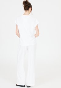 Valkoinen puuvillapaita lyhyillä hihoilla, yhdistettynä löysiin valkoisiin pellavahousuihin. Mallia katsotaan takaa. Mustat kengät näkyvissä.