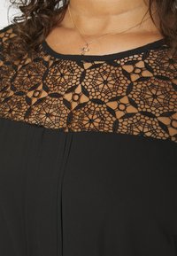 Personne aux cheveux bouclés porte une robe noire avec un empiècement en dentelle florale transparente et un collier en or avec un petit pendentif en forme de croix.
