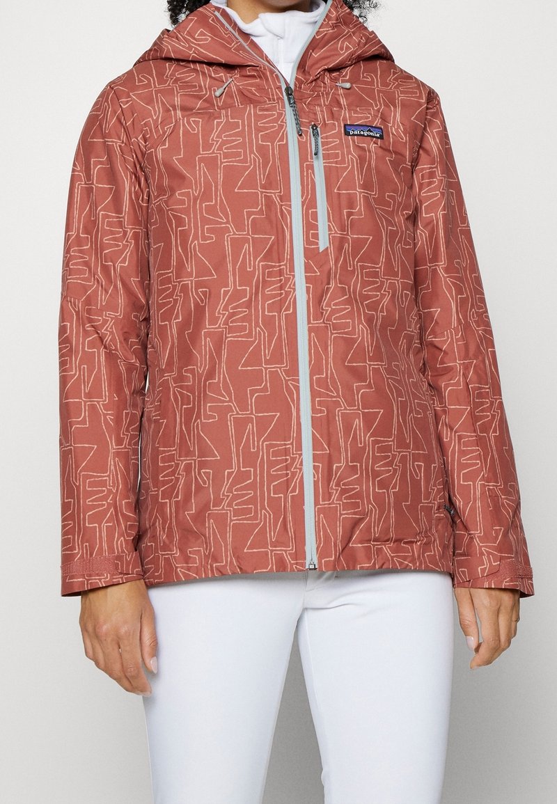 Veste à capuche de couleur rouille à motif, fermeture éclair sur le devant et poche poitrine, portée sur un top blanc et associée à un pantalon blanc.