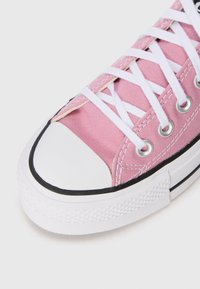 Canvas sneaker with a pink upper, white rubber toe cap, and black accents. Features white laces and silver eyelets. Textured white sole.