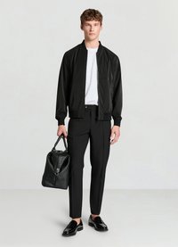 Blouson aviateur noir avec fermeture éclair, associé à un t-shirt blanc, un pantalon noir ajusté et des chaussures noires brillantes. Tenant un sac en cuir noir.