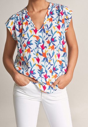 Frau trägt eine ärmellose weiße Bluse mit buntem roten, orangefarbenem, gelbem und blauem Blumenmuster, kombiniert mit weißen Hosen und hat die Hände in den Taschen.