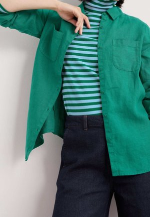 Personne portant une chemise verte à boutons par-dessus un col roulé rayé vert et bleu clair et un jean bleu foncé, avec un bras plié près du visage.