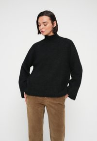 Femme aux cheveux courts et foncés portant un pull à col roulé noir et un pantalon en velours côtelé marron, sur un fond blanc uni.