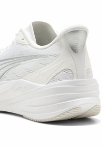 Chaussure de sport blanche avec tige texturée, talon renforcé et design de semelle superposée. Présente une finition lisse et un marquage visible sur le côté.