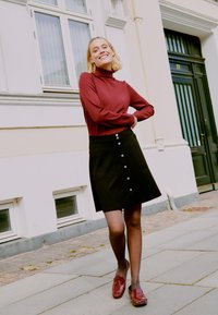 Pull col roulé bordeaux, jupe noire en A avec des boutons sur le devant et mocassins bordeaux brillants. Fond uni avec une fenêtre et une porte.
