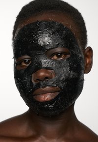 Svart ansiktsmask som täcker ansiktet och framhäver den släta huden under. Masken har en blank yta med utskärningar för ögon och mun.