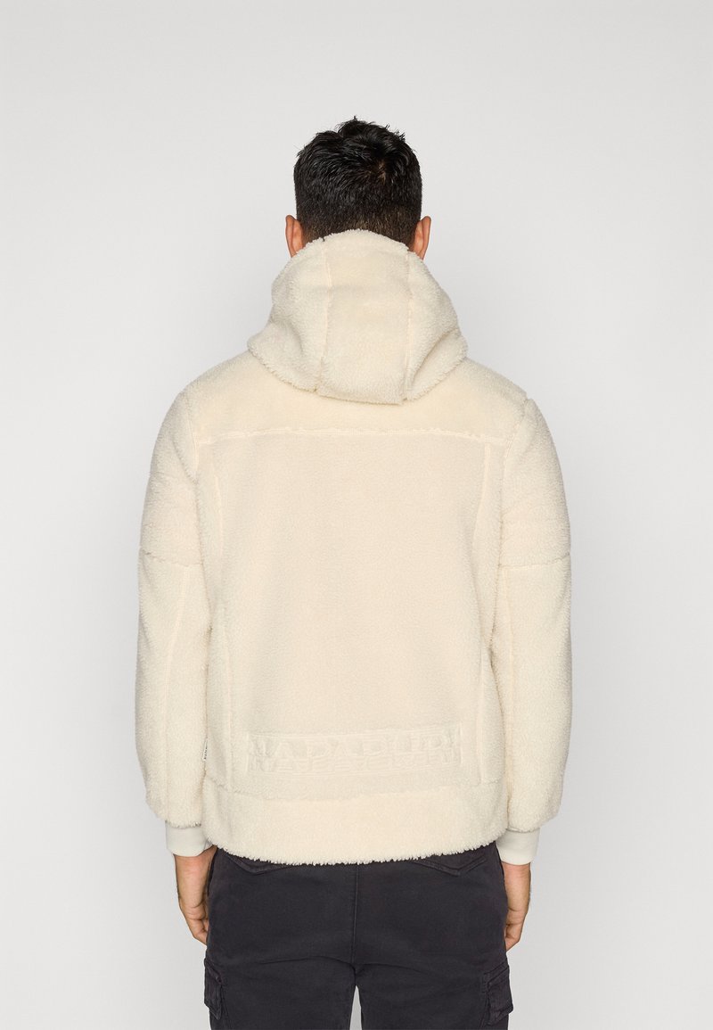 Mann in einem cremefarbenen Fleece-Hoodie mit herabgelassener Kapuze, von hinten gesehen, kombiniert mit dunklen Cargo-Hosen vor einem schlichten Hintergrund.