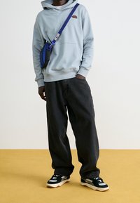 Felpa con cappuccio azzurro chiaro con tasca frontale e logo, abbinata a jeans neri larghi e sneaker nere e bianche. Borsa a tracolla blu aggiunge colore.