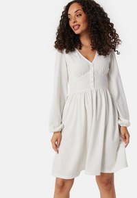 Abito bianco a maniche lunghe con scollatura a V, dettagli in bottone, tessuto testurizzato e vita elasticizzata, che crea una silhouette fluida.