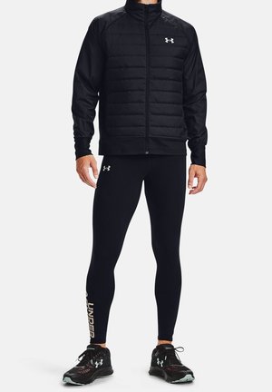 Mężczyzna w czarnej pikowanej kurtce, czarnych dopasowanych legginsach z logo marki na nodze oraz czarnych butach sportowych z logo.