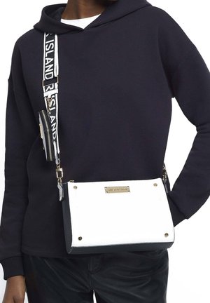 Persoon draagt een zwarte hoodie en zwarte broek met een witte en zwarte crossbodytas met het label "River Island" en bijpassende riem.